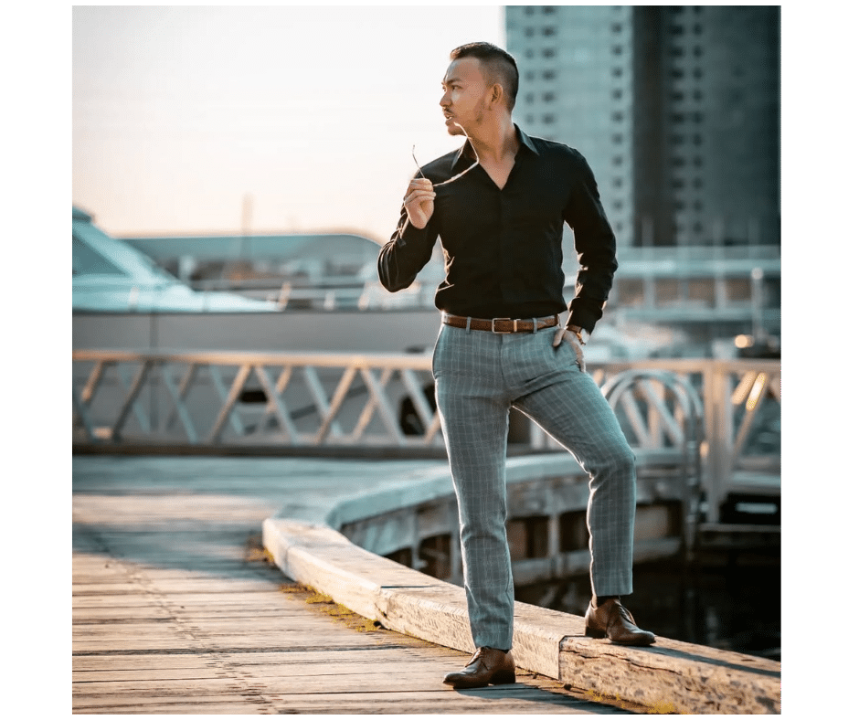 A man in a black shirt and plaid pants stands on a dock, holding sunglasses and looking to the side, with boats and buildings in the background at sunset.