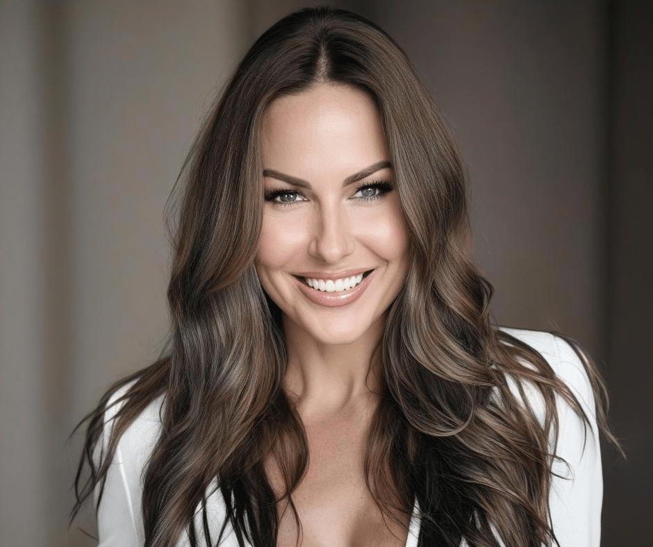 A woman with long, wavy brown hair and light eyes smiles at the camera, wearing a white top. The background is softly blurred.