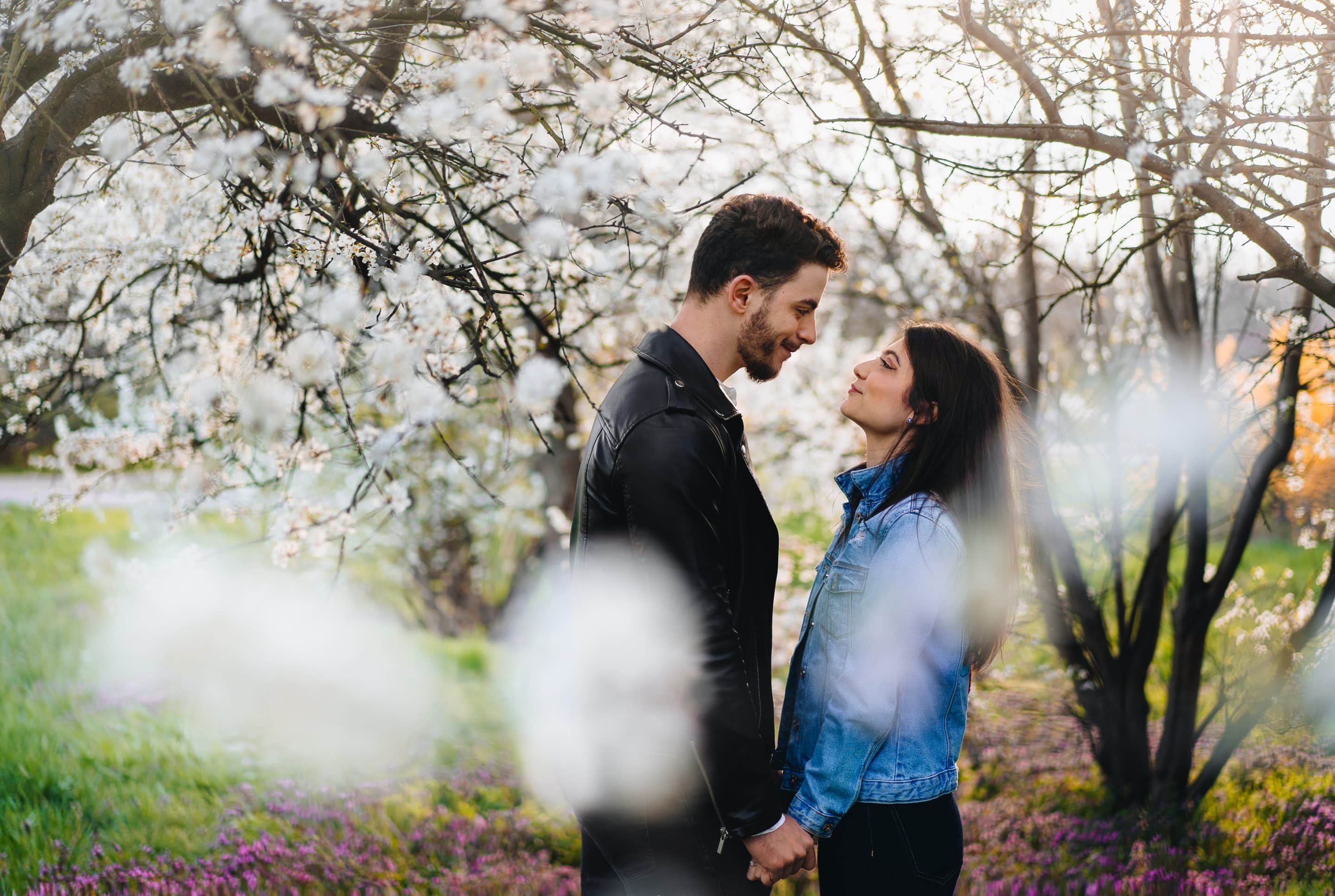A couple stands close together, holding hands and gazing into each other's eyes under blooming cherry blossom trees in a sunlit park.
