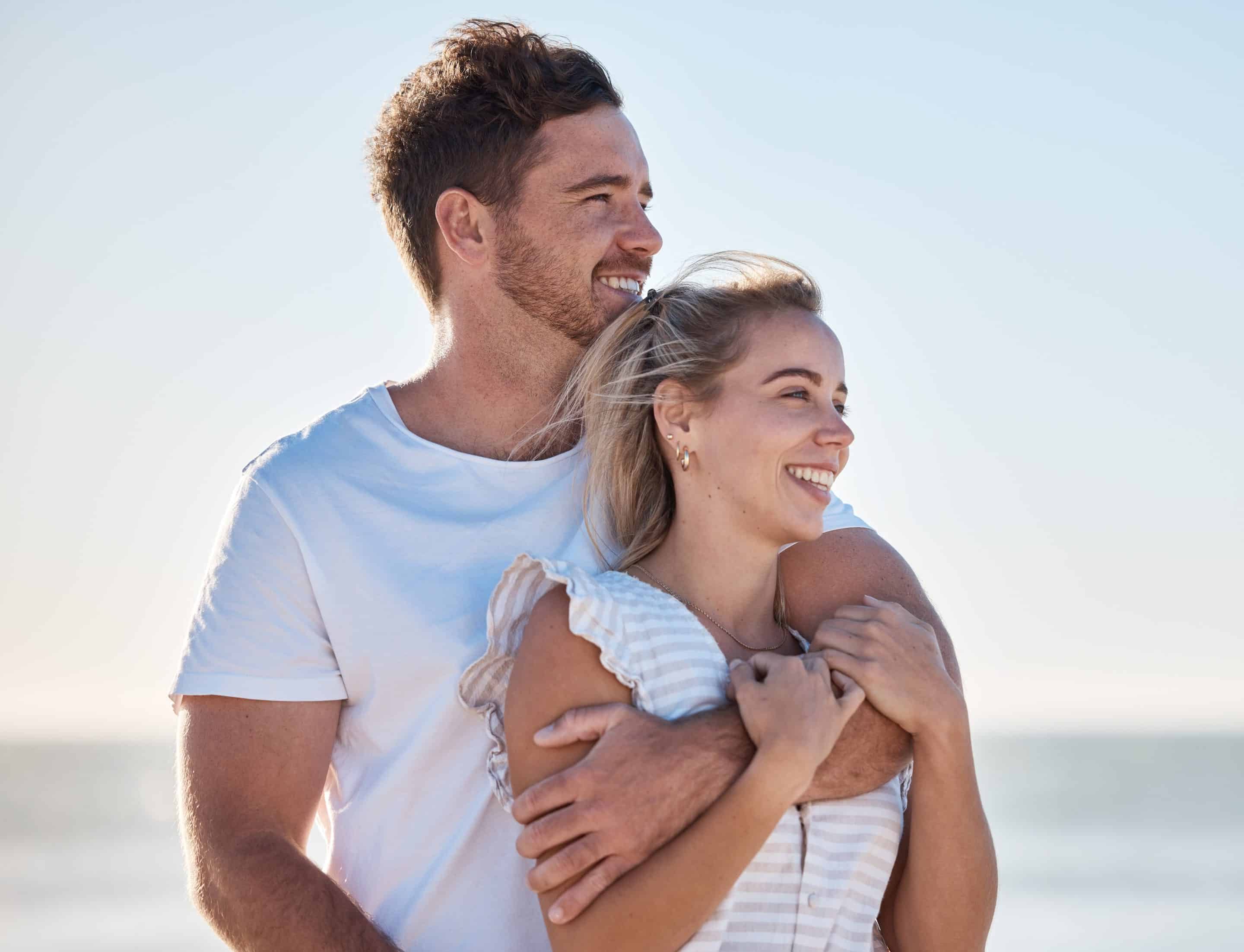 A smiling couple stands on a sunny beach. The man, wearing a white t-shirt, hugs the woman from behind. She is wearing a light striped dress, and both look happy and relaxed with the ocean in the background.