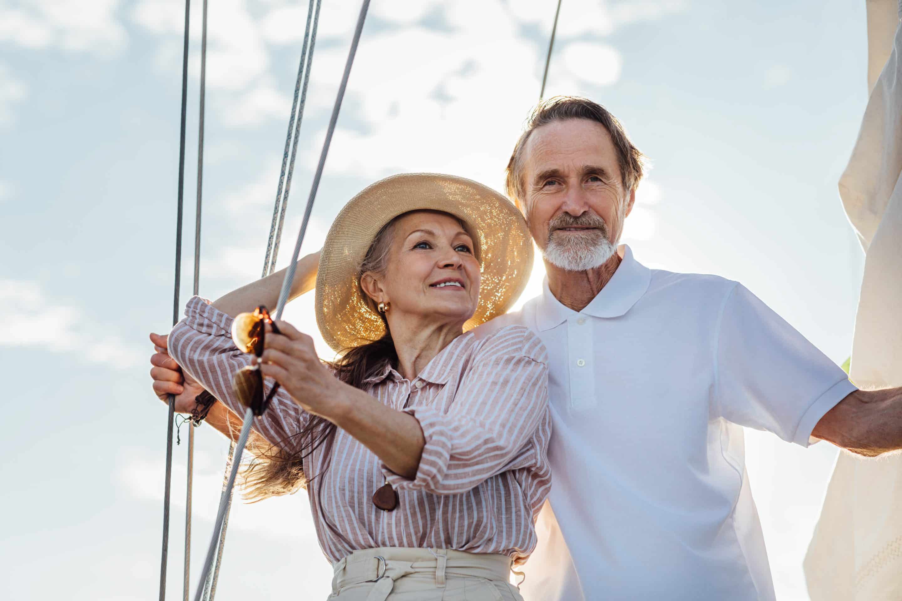 An older couple enjoys sailing on a sunny day. The woman wears a straw hat and striped shirt while holding a rope; the man, in a white polo, stands beside her smiling, with bright sky and sailboat rigging in the background.