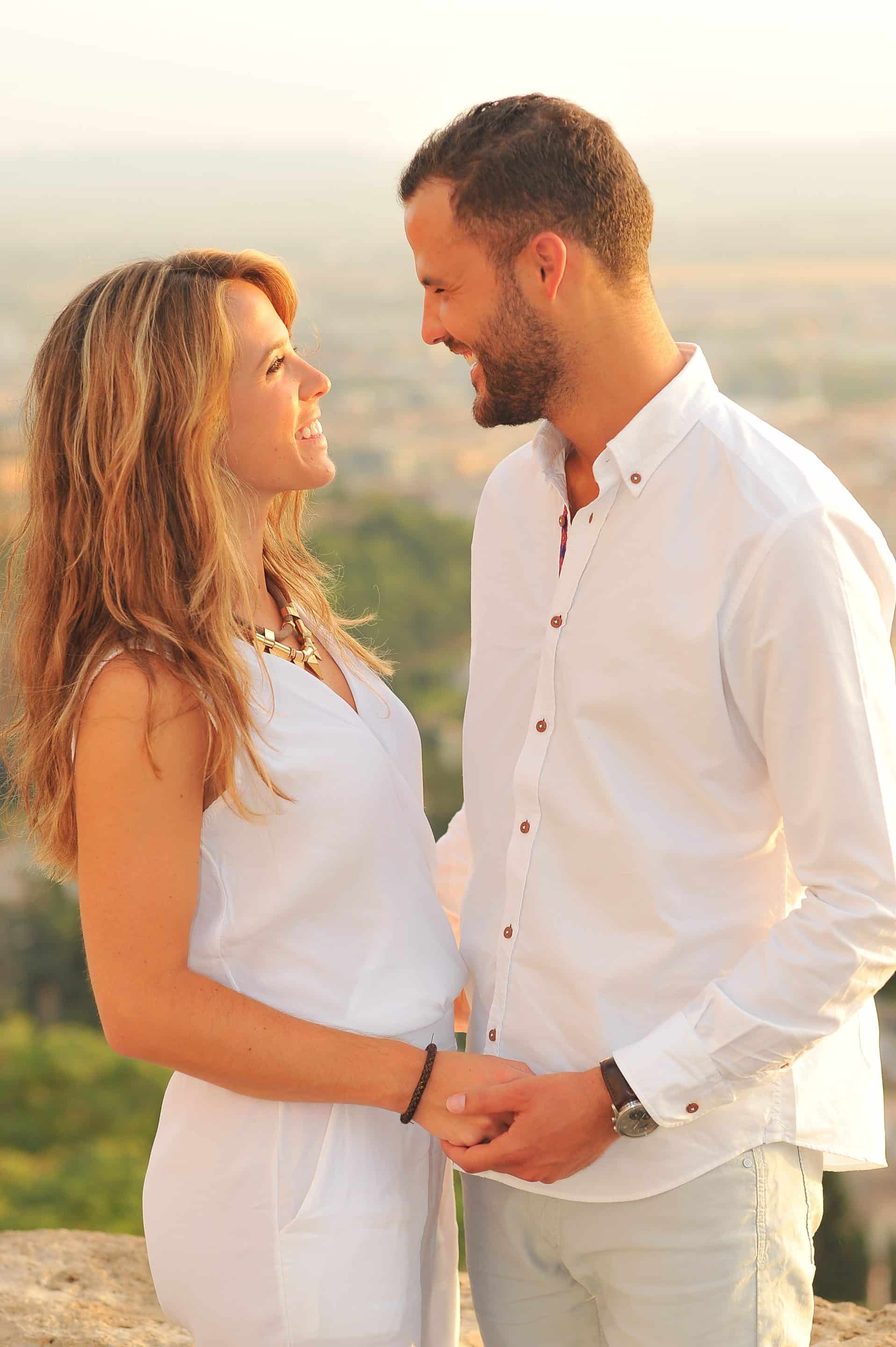 A couple dressed in white stands outdoors, smiling and holding hands while gazing into each other's eyes. The background is blurred with greenery and a soft, warm light.