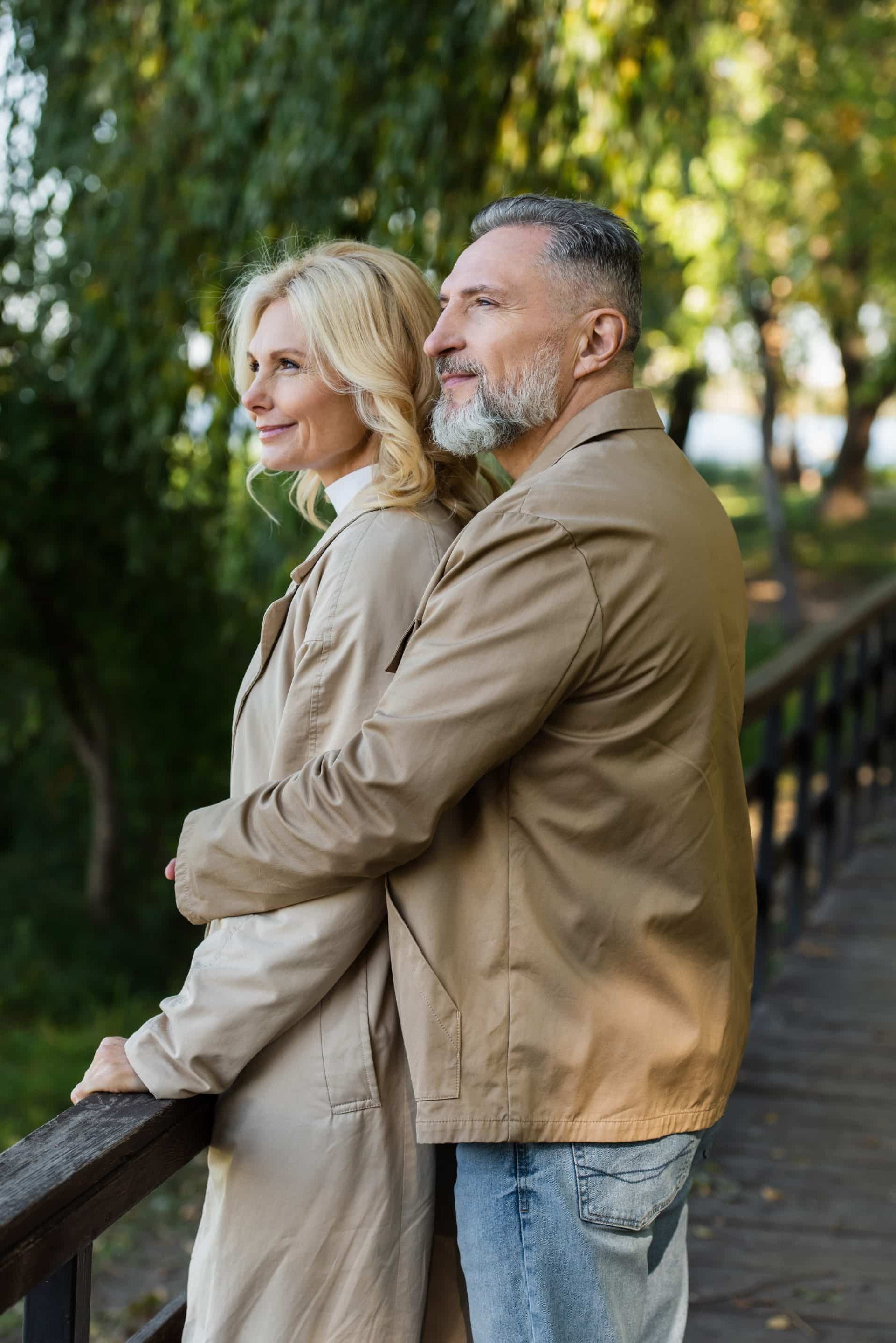 An older couple stands close together on a wooden bridge, both wearing beige jackets. The man embraces the woman from behind as they look into the distance, surrounded by green trees and natural sunlight.