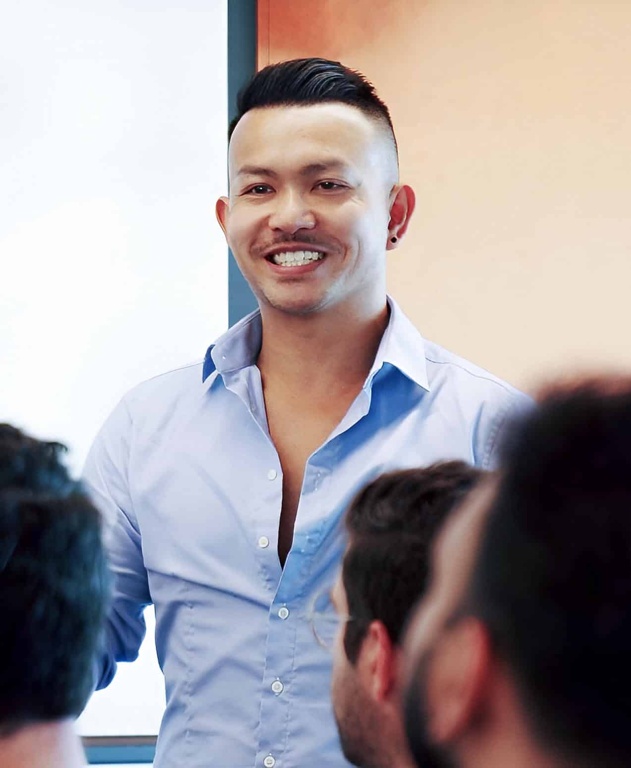 A man wearing a light blue shirt is smiling and speaking to a group of people in a brightly lit room. The focus is on him, while the audience is blurred in the foreground.