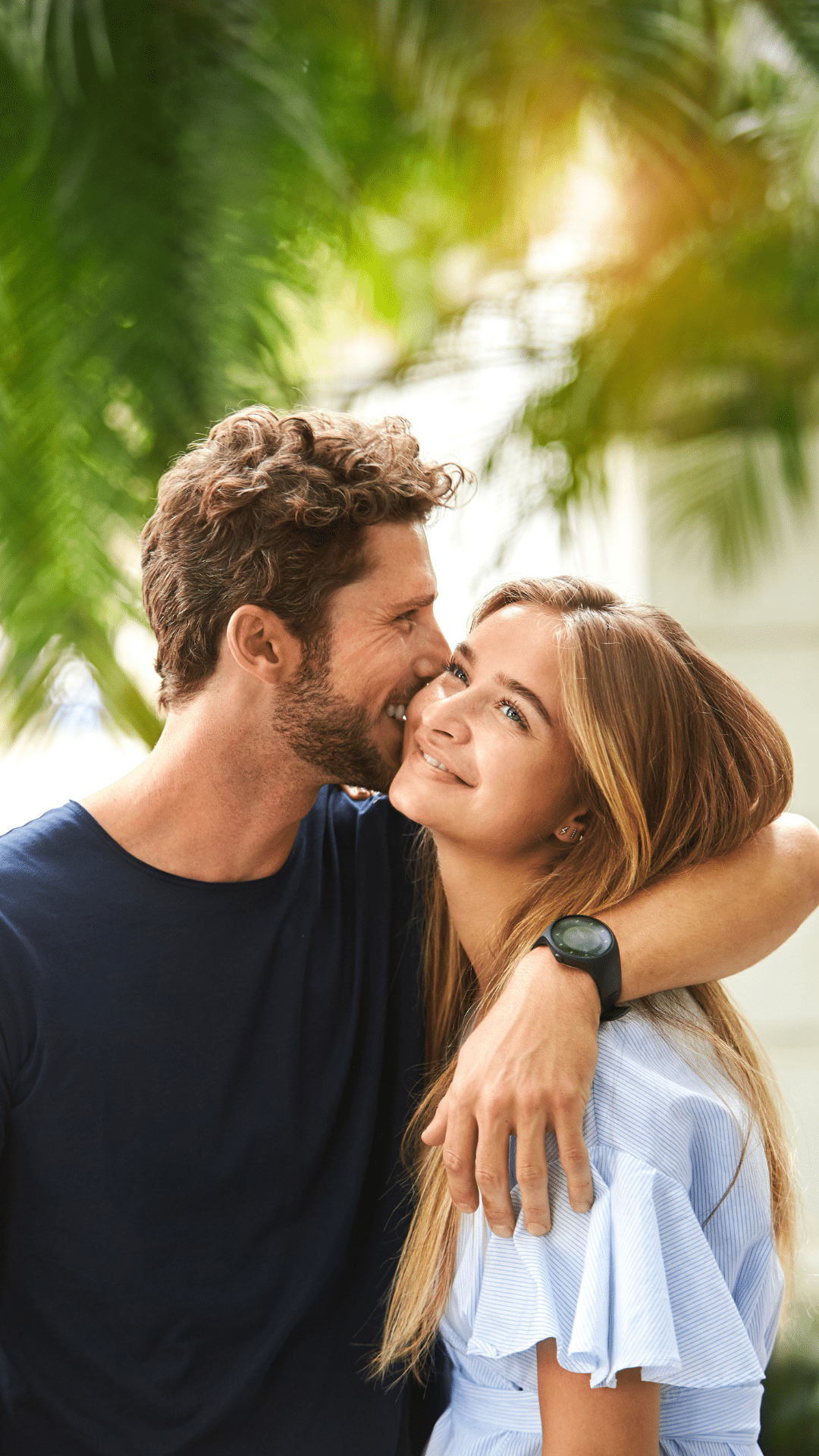 A man with a beard hugs a smiling woman from the side, whispering into her ear. They stand outdoors in front of green, leafy foliage, both wearing casual clothes and looking happy.
