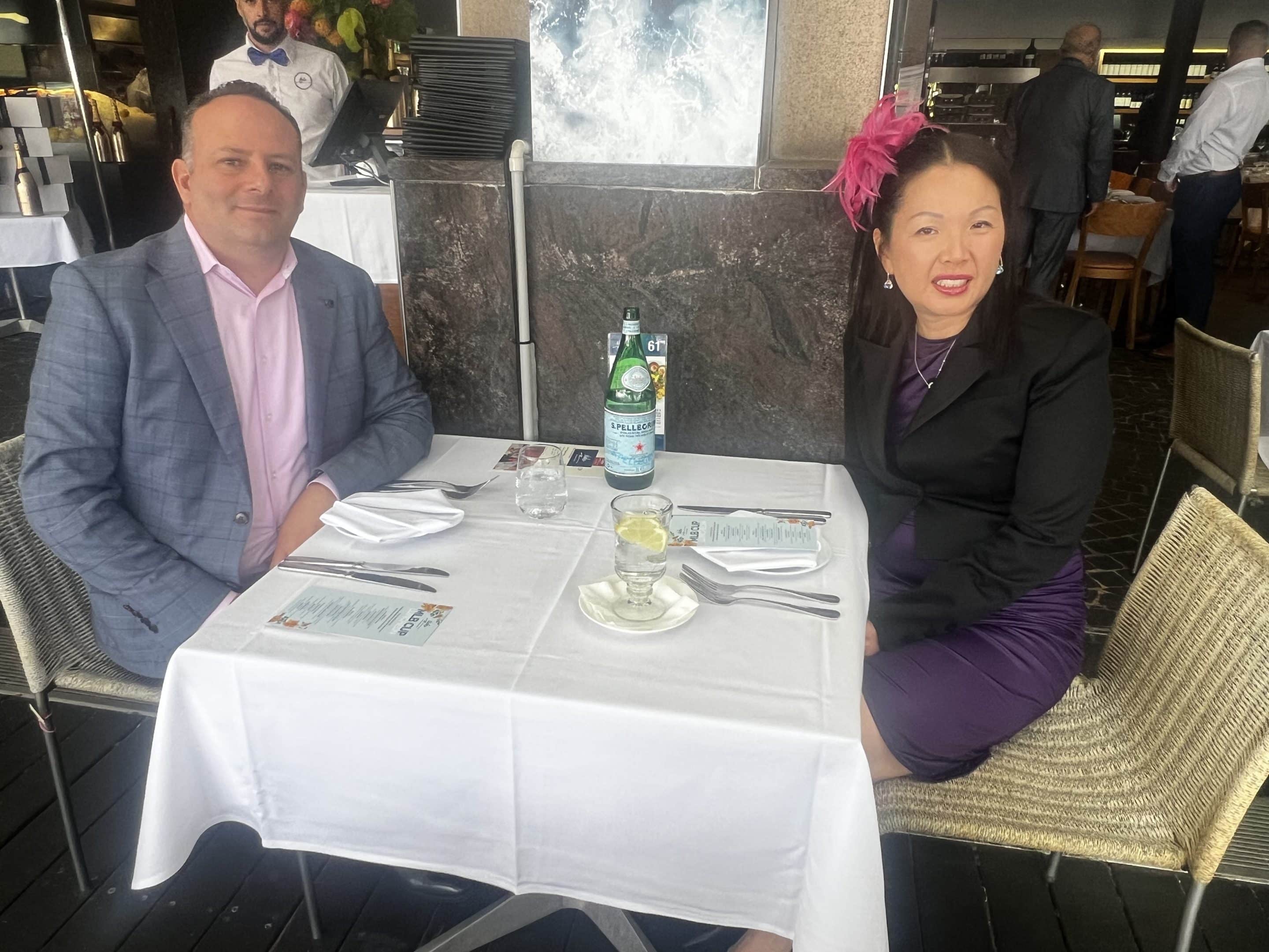 A man in a light blue suit and a woman in a purple dress with a pink fascinator are seated at a white tablecloth-covered table in a restaurant, smiling at the camera. Bottled water and menus are on the table.