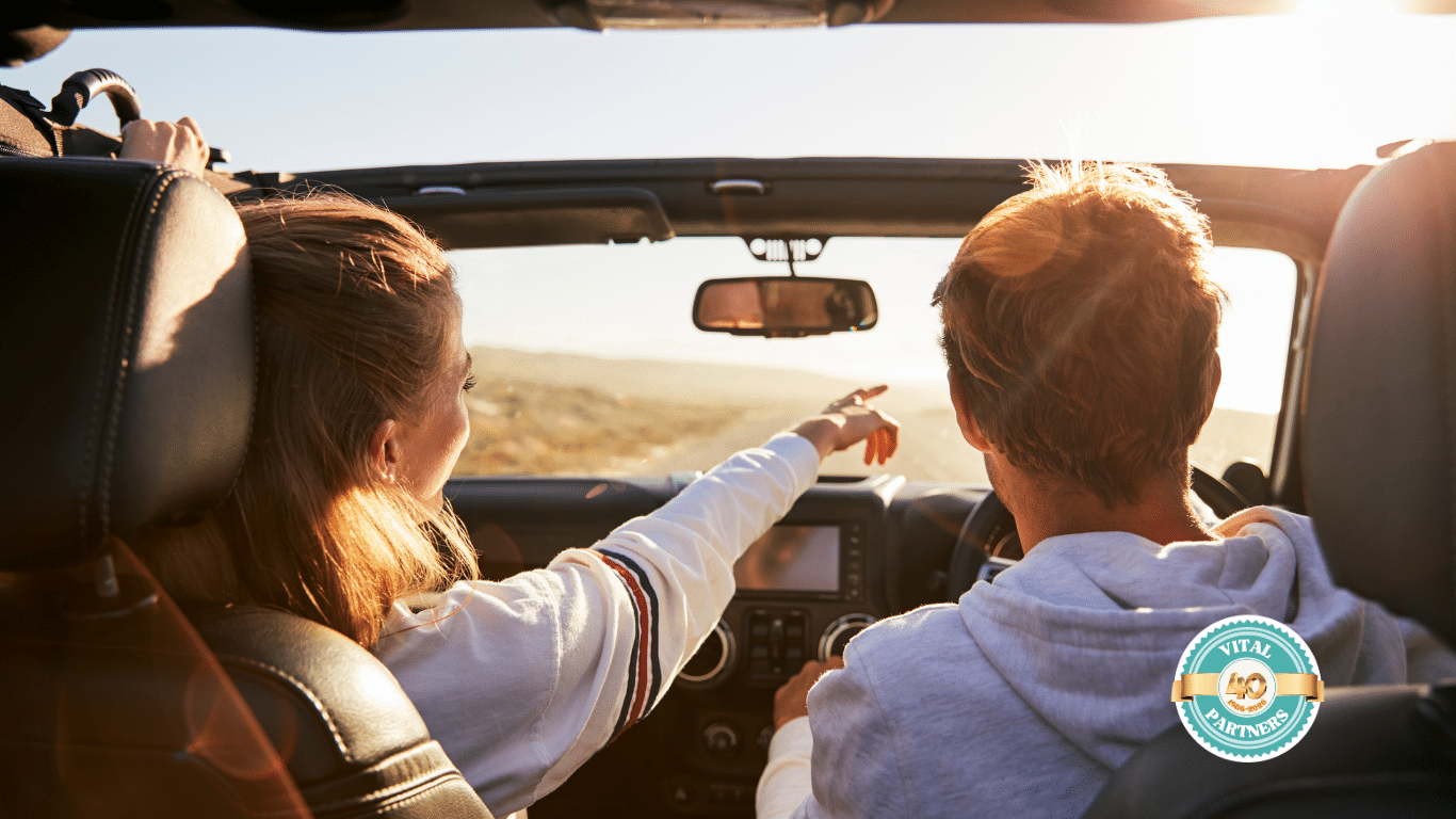 Two people drive in a convertible with the top down at sunset. The passenger points ahead, smiling, while the driver looks forward. Sunlight streams in, creating a warm atmosphere. A circular “Vital Partners” logo is in the lower right corner.