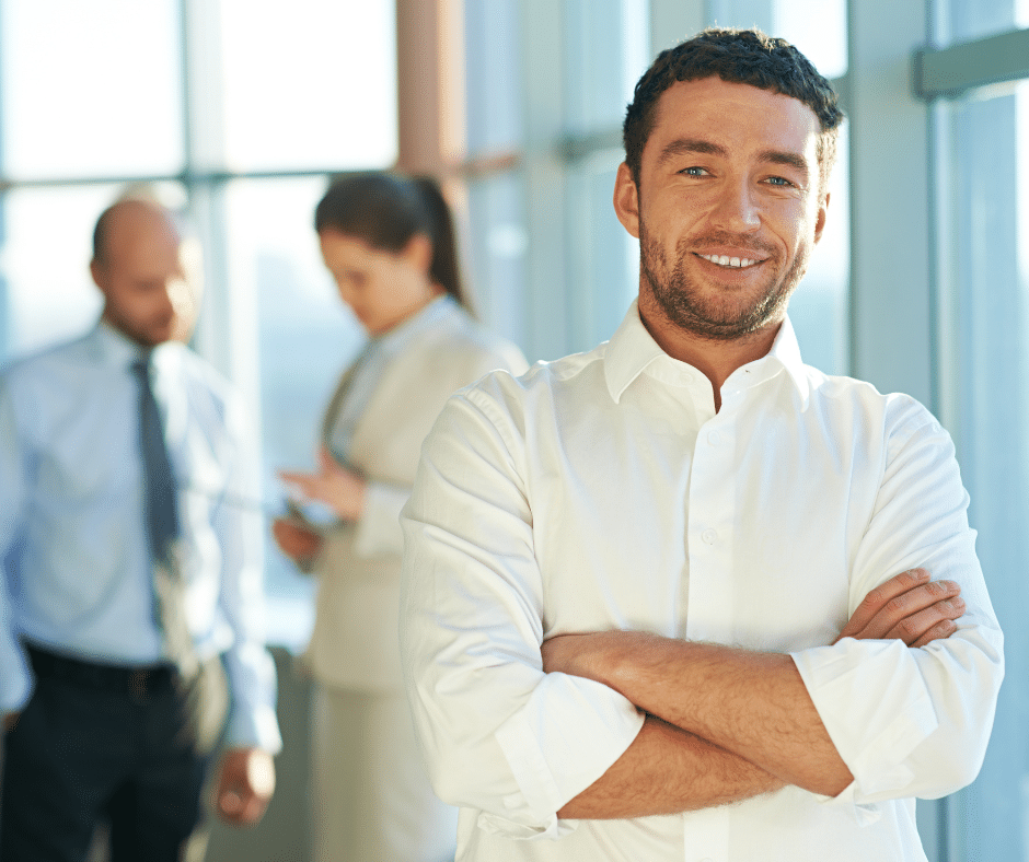 A man in a white shirt stands with arms crossed and smiles at the camera, while two colleagues in business attire talk in the blurred background near large windows.