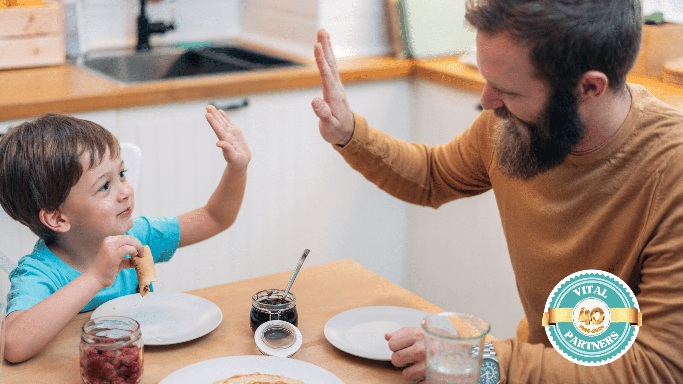 A smiling man and child sit at a kitchen table, giving each other a high-five. The child holds a rolled pancake, with jars of jam and syrup on the table. The "Vital Partners" logo appears in the bottom right corner.