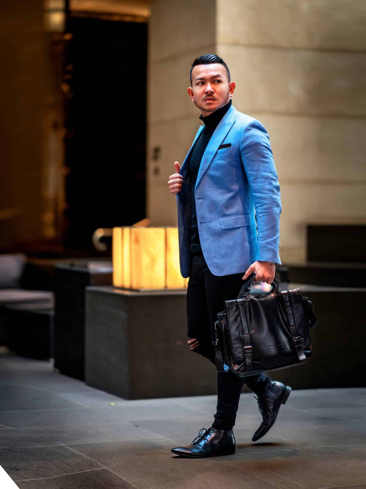 A man in a light blue blazer, black turtleneck, and black pants walks indoors, holding a black leather bag and giving a thumbs-up gesture. The background features a modern, softly lit interior.