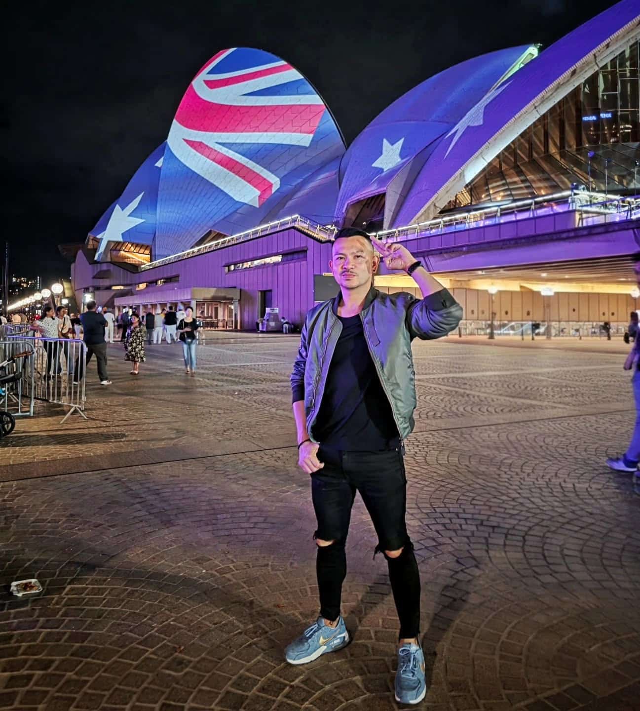 A man poses at night in front of the Sydney Opera House, which is illuminated with the Australian flag. The area is well-lit, with several people and barriers visible in the background.