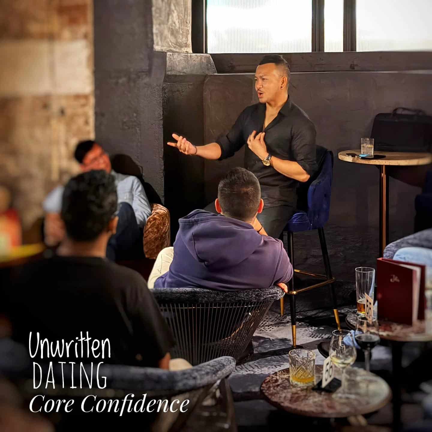 A man in black speaks confidently to a small group seated in a dimly lit lounge. Text reads: "Unwritten DATING Core Confidence." Drinks and notebooks are on the table in the foreground.