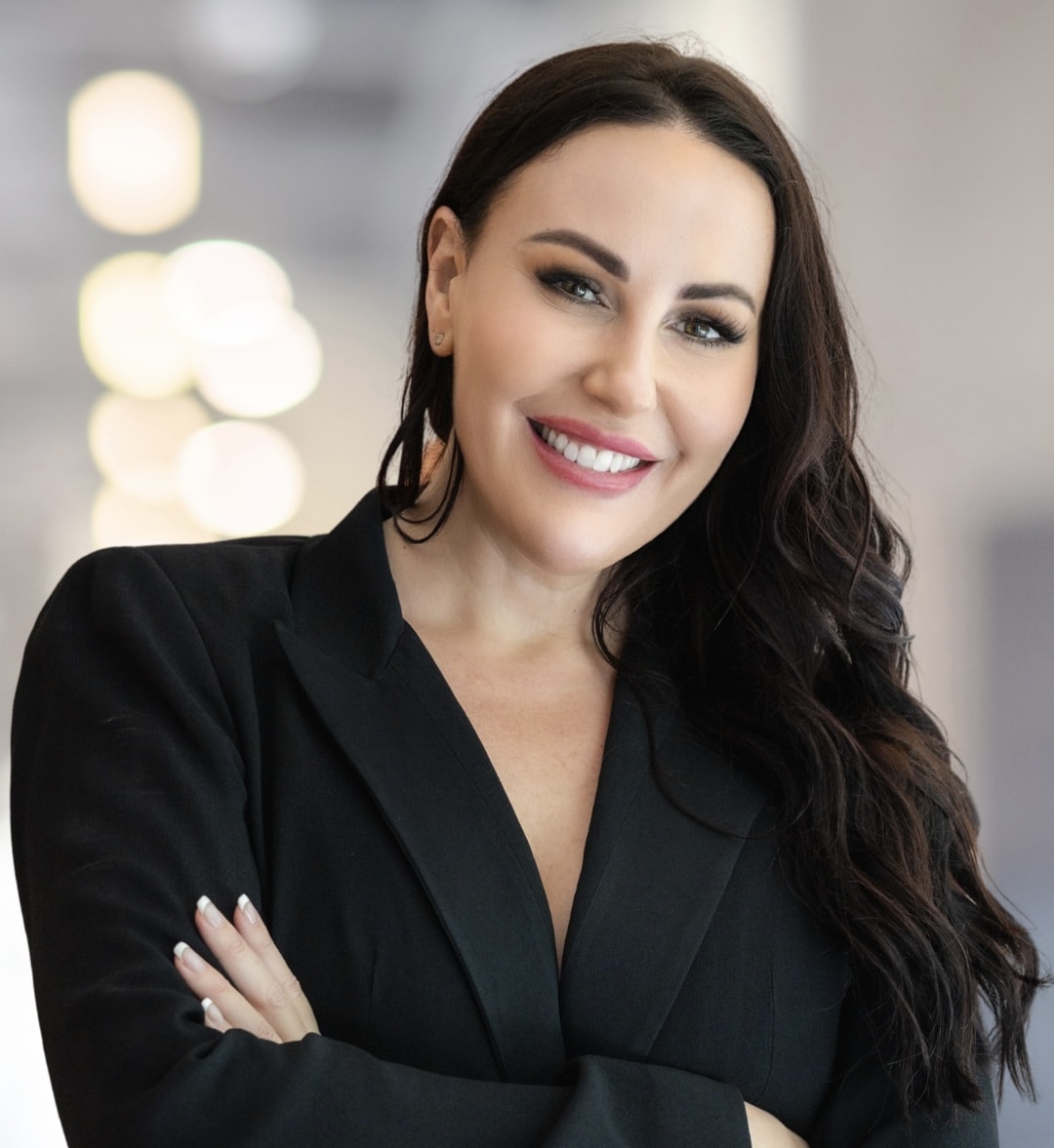 Smiling woman with long dark hair wearing a black blazer, standing with arms crossed in a bright, blurred indoor setting.