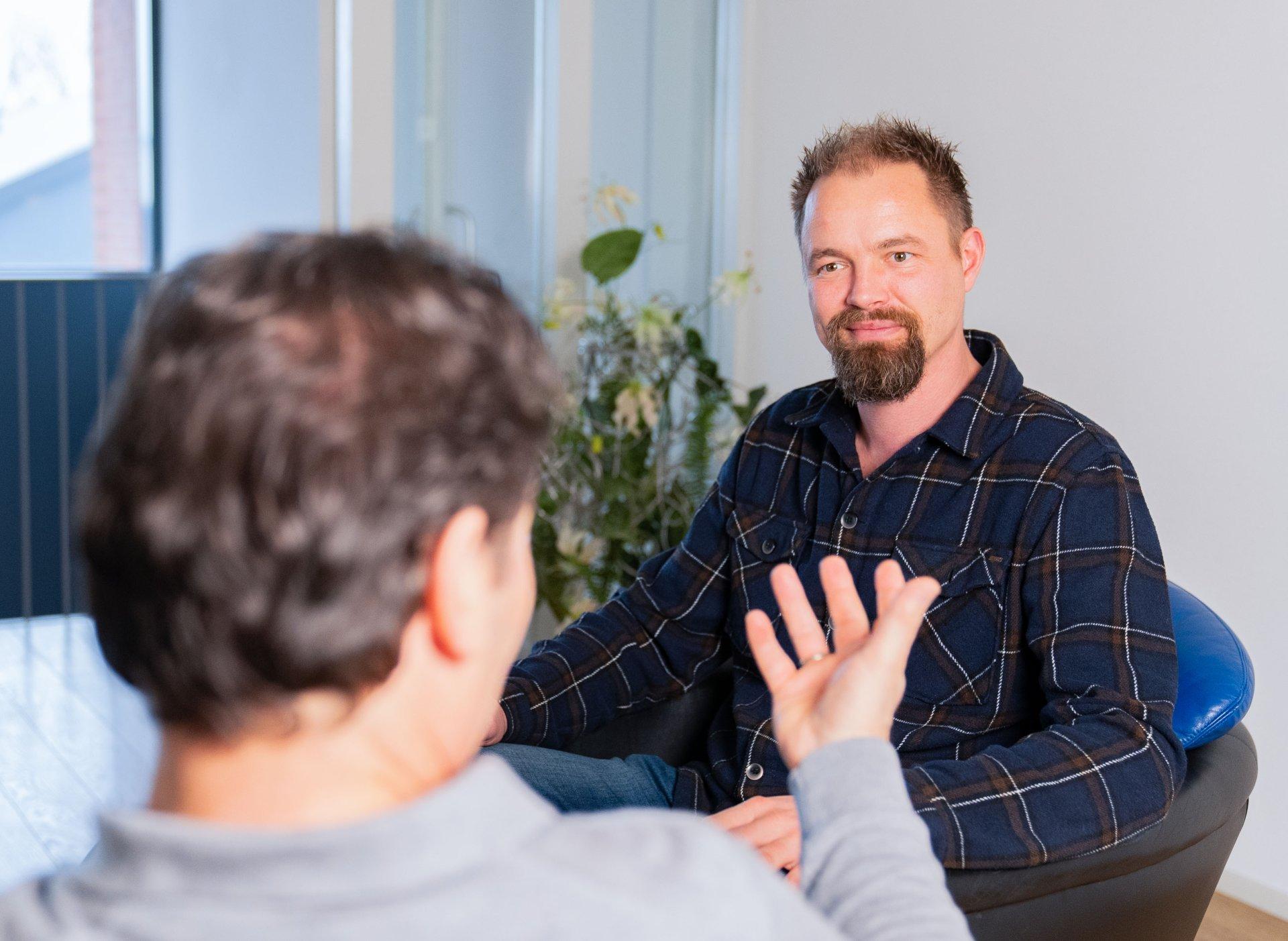 Two men are sitting and talking indoors. One man faces the camera, smiling and listening, while the other is turned away, gesturing with his hand as he speaks. There is a plant in the background.