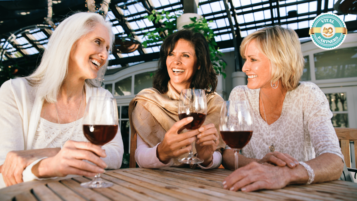Three women sit at a wooden table, smiling and enjoying glasses of wine together in a bright, glass-roofed setting with plants. A circular "Vital Partners 50" logo is visible in the upper right corner.