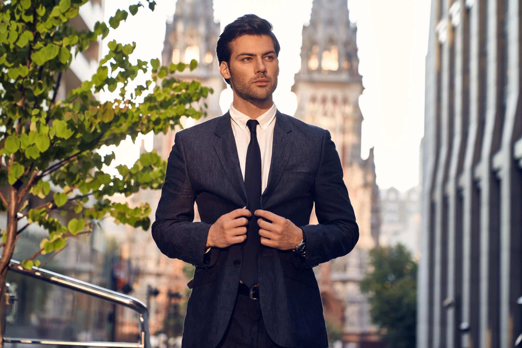 A man in a dark suit and tie stands outside in an urban setting, adjusting his jacket. Tall, gothic-style buildings and a leafy tree are visible in the background.