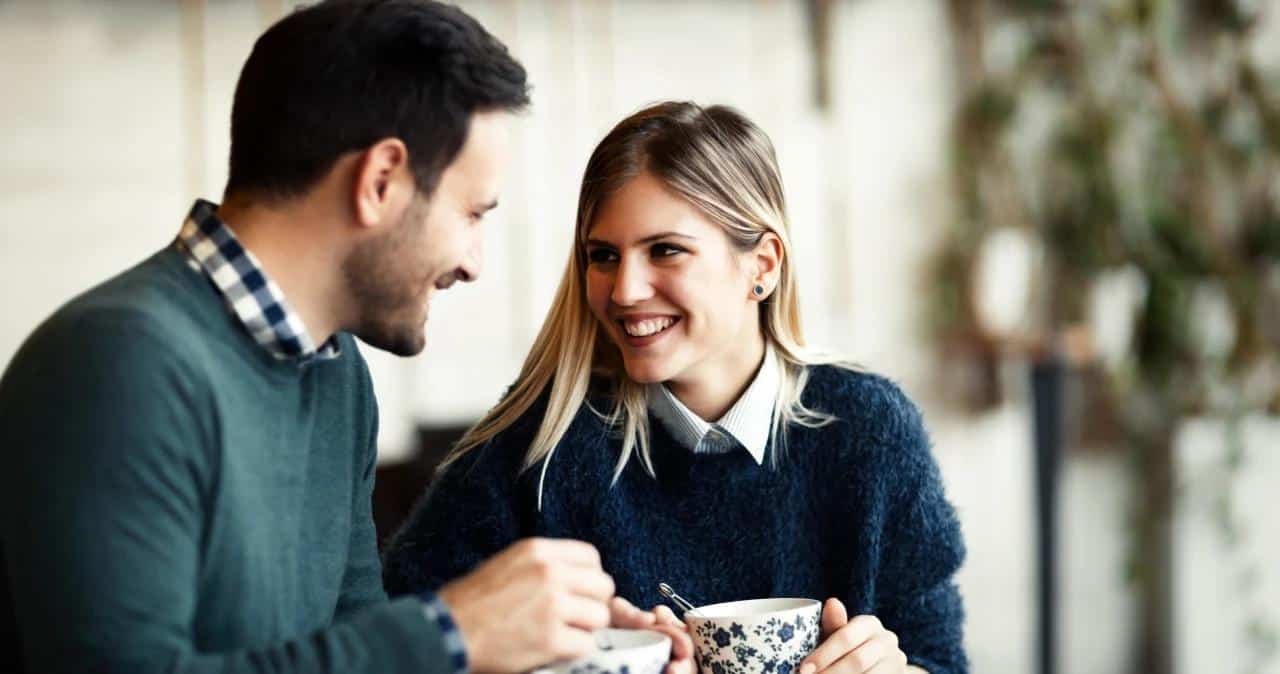 A man and a woman sit together at a table, smiling warmly at each other while holding mugs. The setting appears to be a cozy indoor café with a blurred green background.