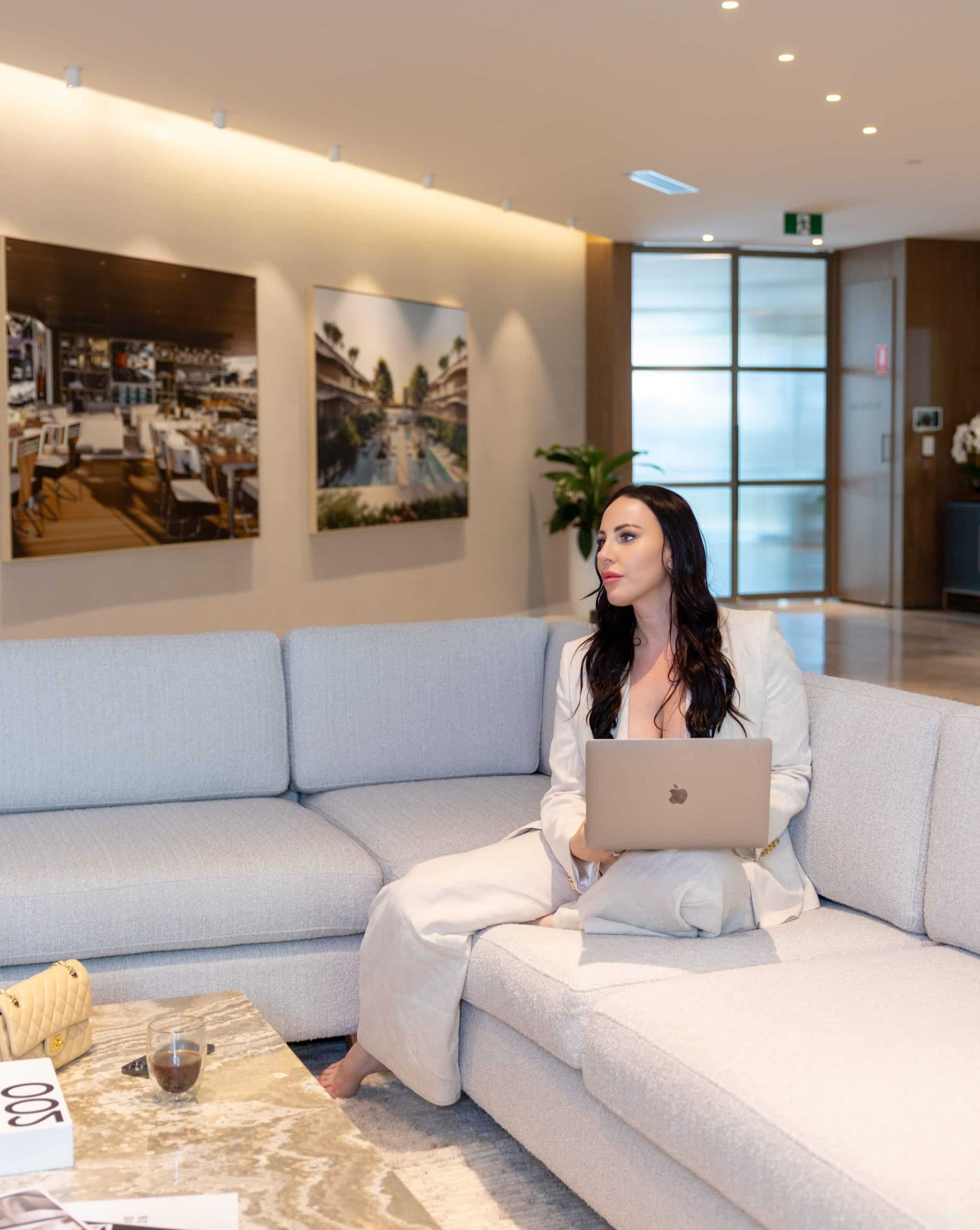 A woman in a light-colored suit sits on a modern gray sectional sofa with a laptop on her lap in a stylish, well-lit office lounge area, with wall art and a coffee table nearby.