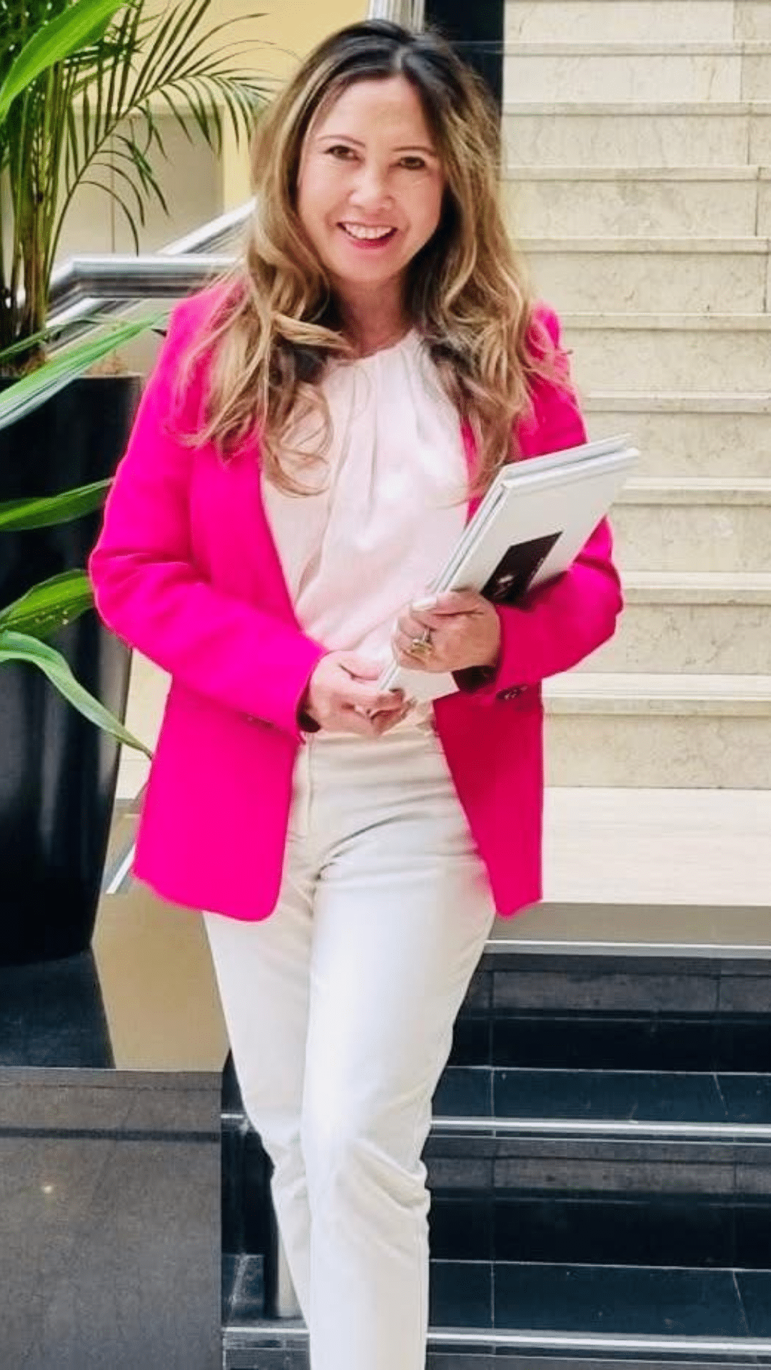 A woman with long, wavy hair wearing a bright pink blazer and white pants stands indoors holding a laptop and papers, smiling in front of a staircase and a potted plant.