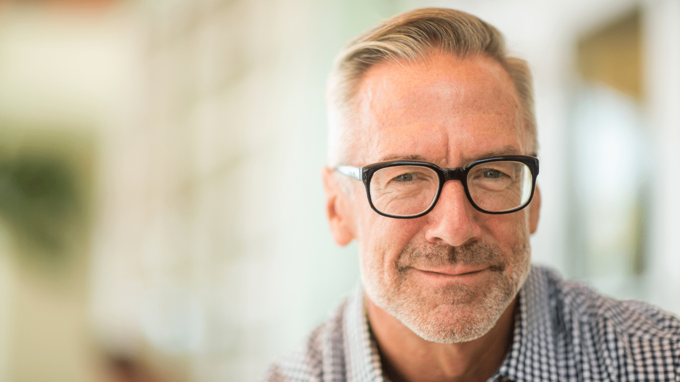 A middle-aged man with gray hair, a beard, and black-rimmed glasses smiles slightly while wearing a checked shirt. The background is softly blurred, focusing attention on his face.