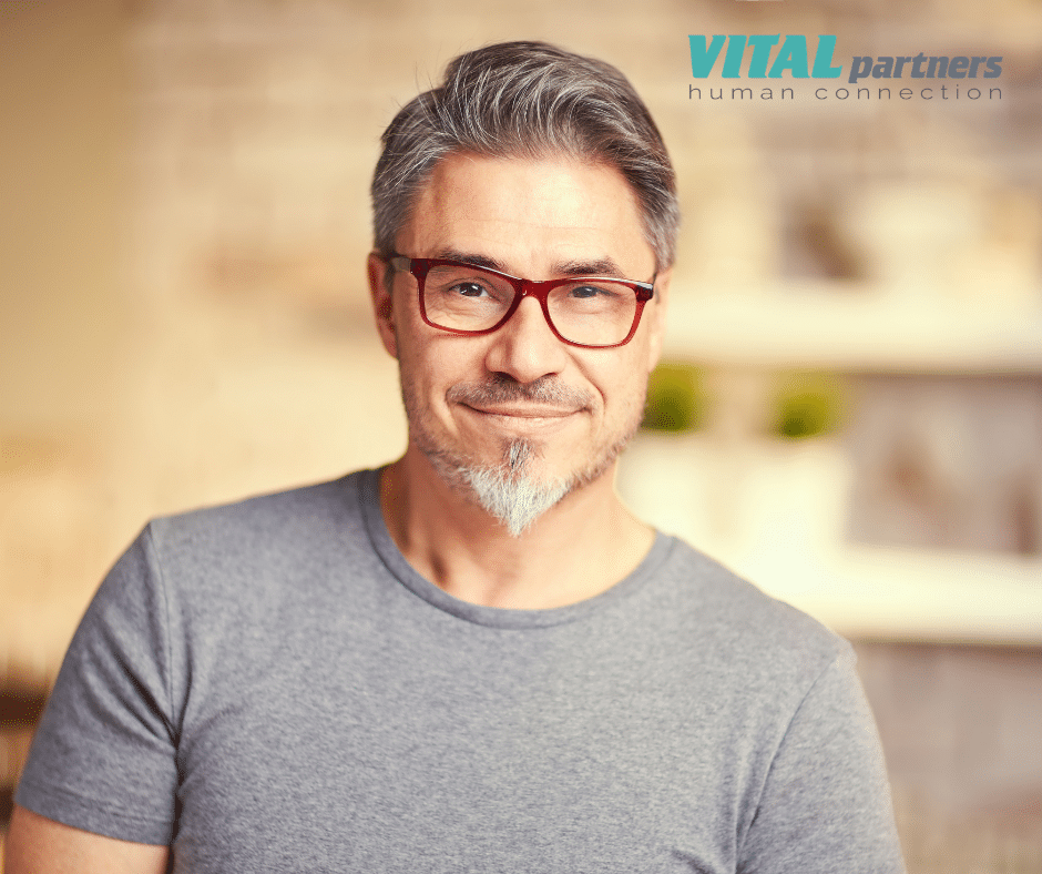 Smiling man with gray hair, glasses, and short beard wearing a gray t-shirt. Blurred indoor background. “VITAL partners human connection” logo appears in the top right corner.