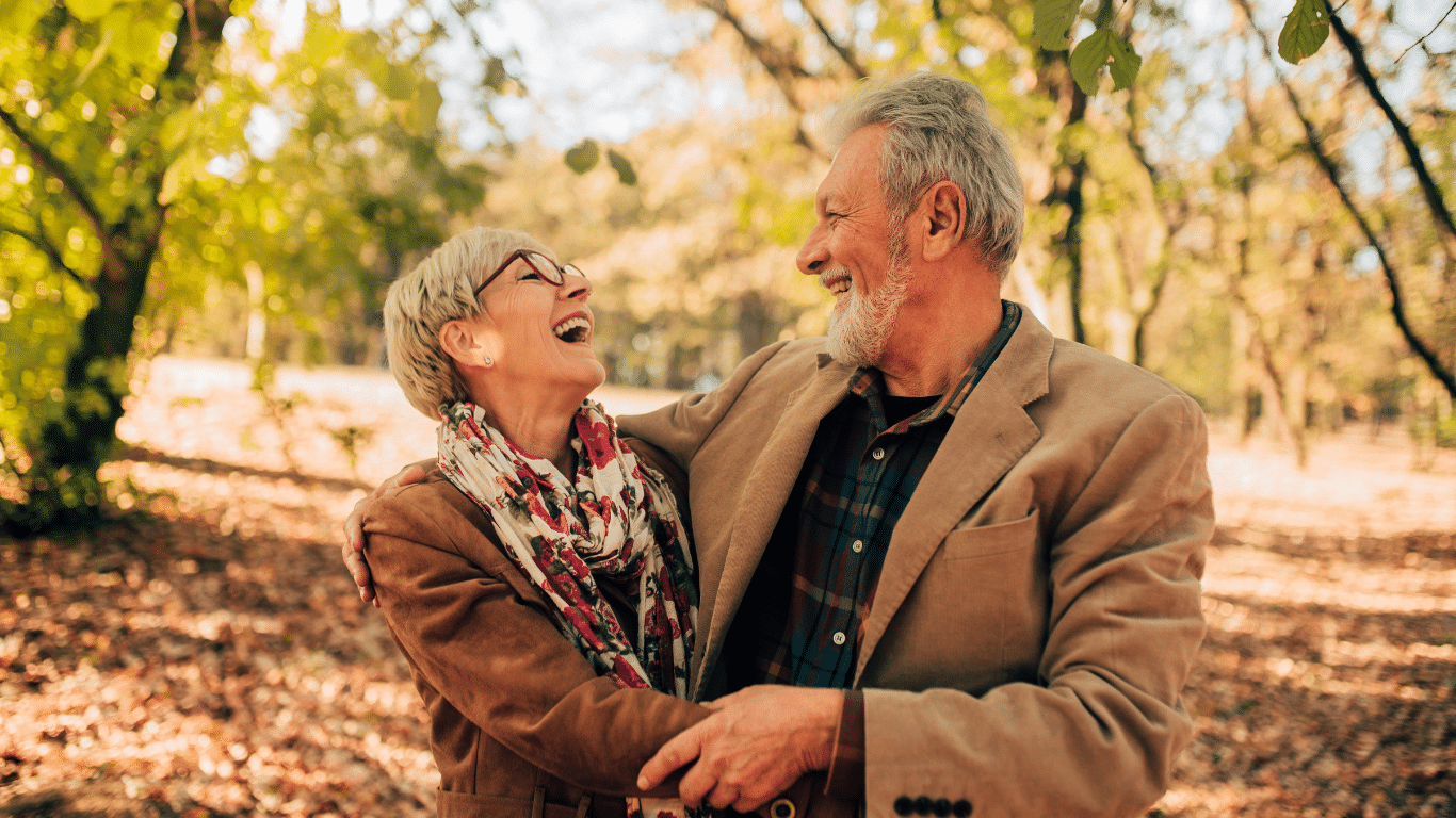 An older couple laughs while embracing in a wooded park during autumn.